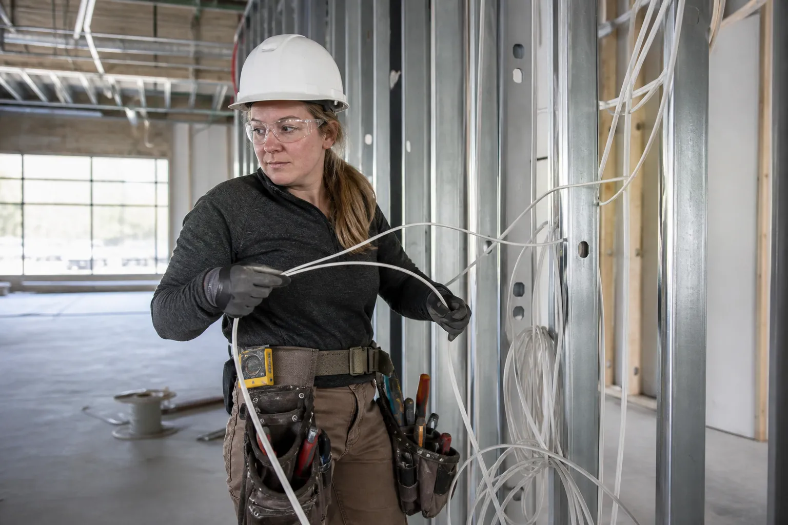 An electrician in a white CSA Type 1 Class E hard hat, arc-rated FR clothing under a Class 2 hi-vis vest, safety glasses and CSA electrical-hazard boots pulling cable in a bright commercial build.