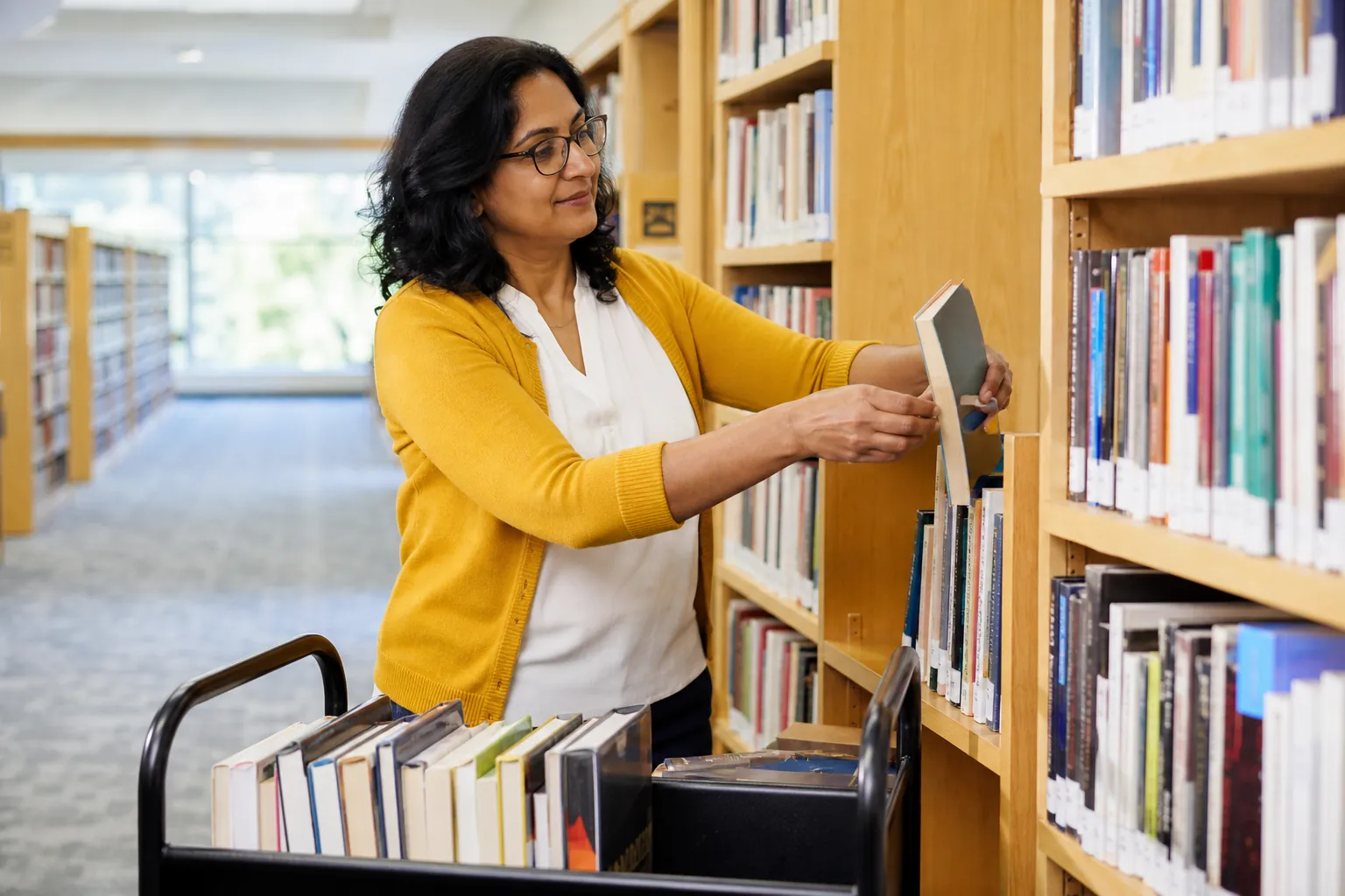 A public-library worker reshelving books from a rolling cart in a bright branch.